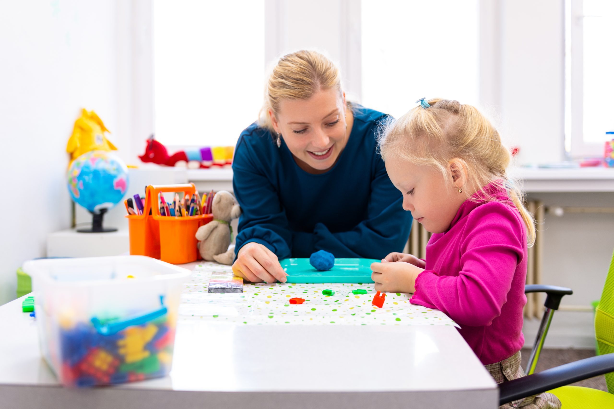 A smiling woman helping a child with a craft activity in a brightly lit, colourful room.